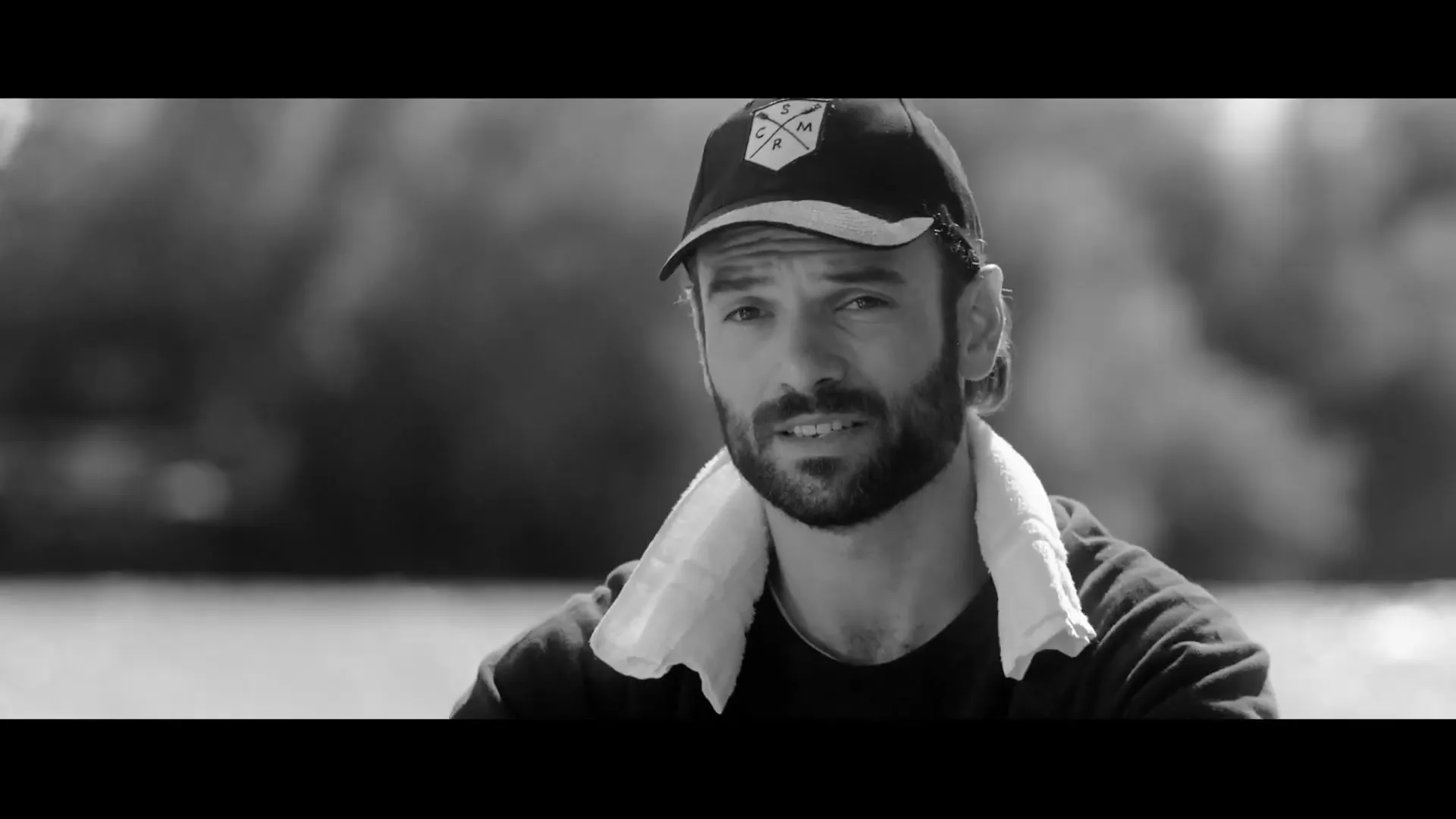 A man with a beard and a cap, wearing a towel around his neck, looks directly at the camera in a medium shot, outdoors by a body of water.