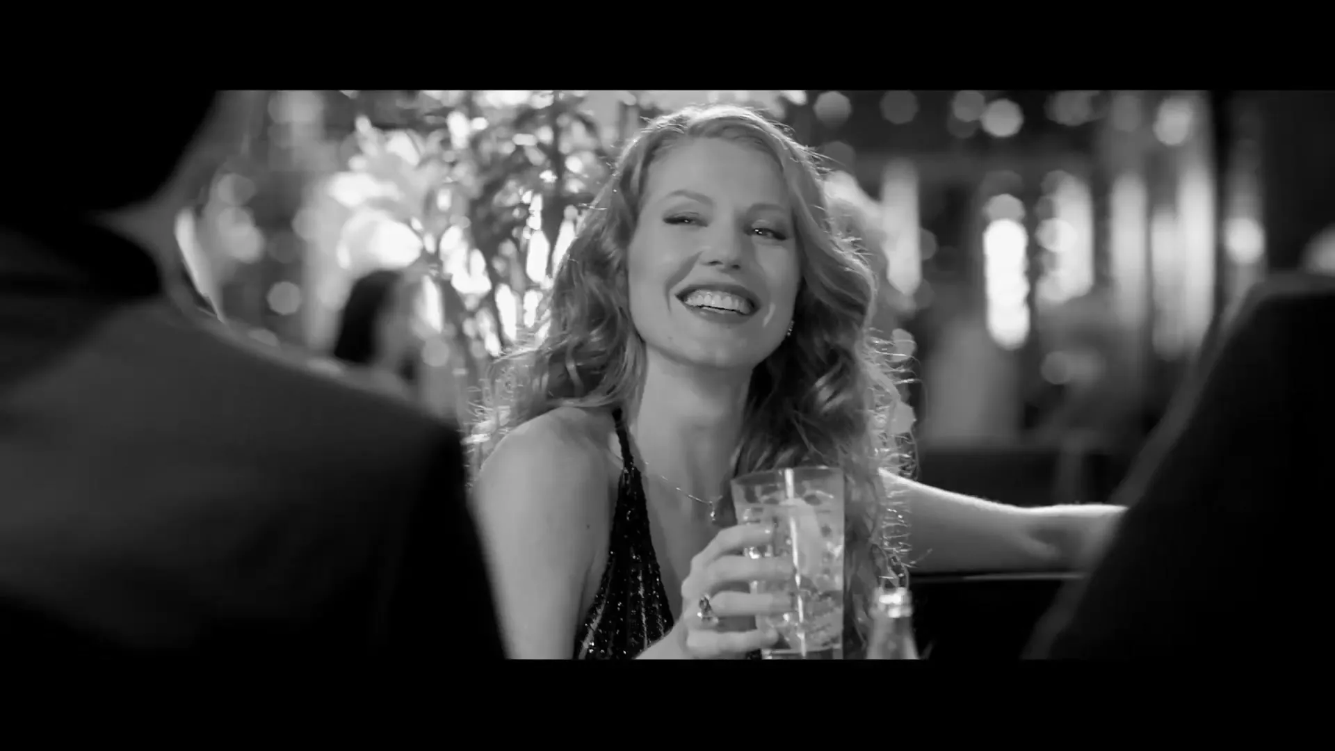 A beautiful woman with long curly hair, wearing a sparkly dress, smiles while holding a drink in a bar, in black and white.