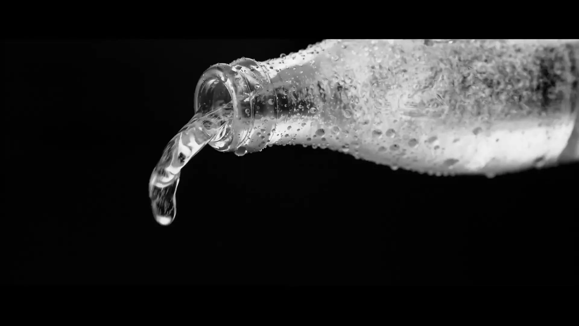 A clear glass bottle, covered in condensation, pours a stream of sparkling water against a black background.