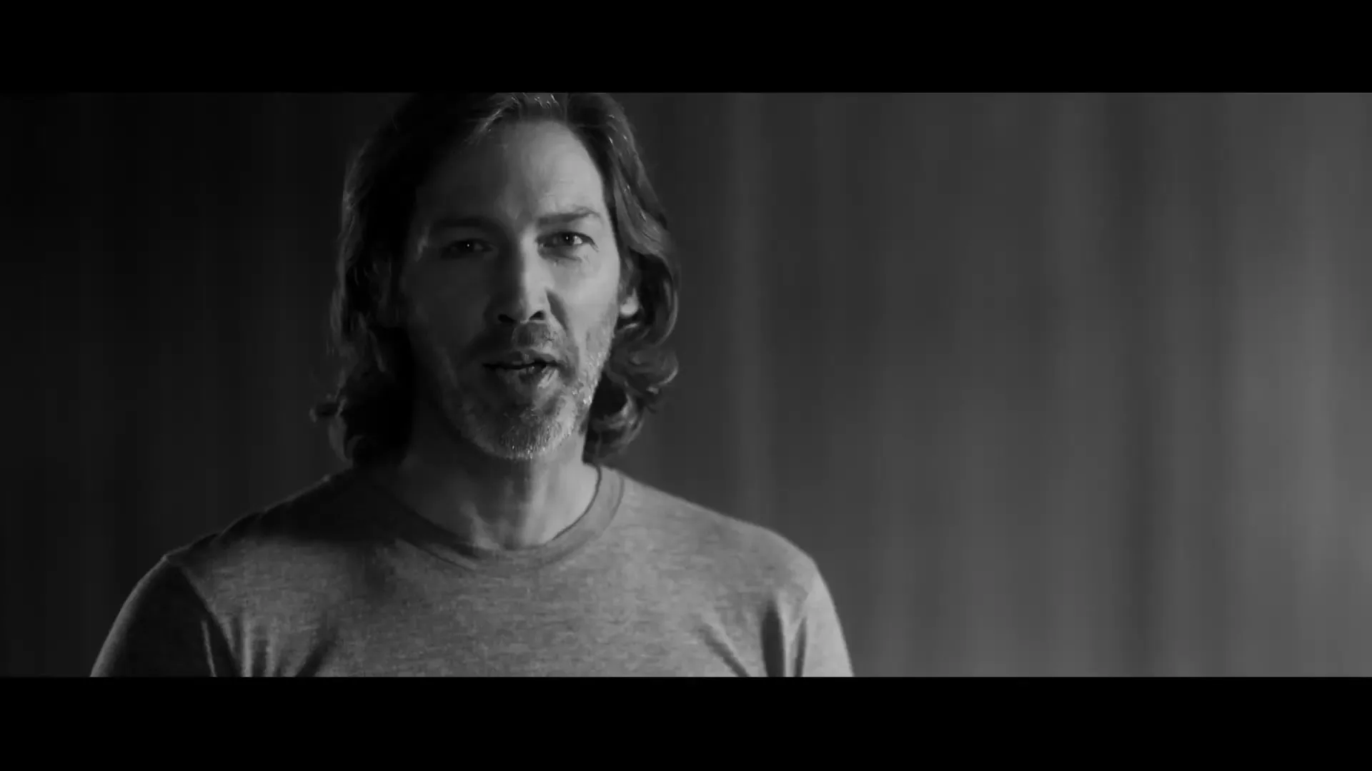 A man with long hair and a beard, wearing a t-shirt, looks directly at the camera and speaks in a black and white shot.