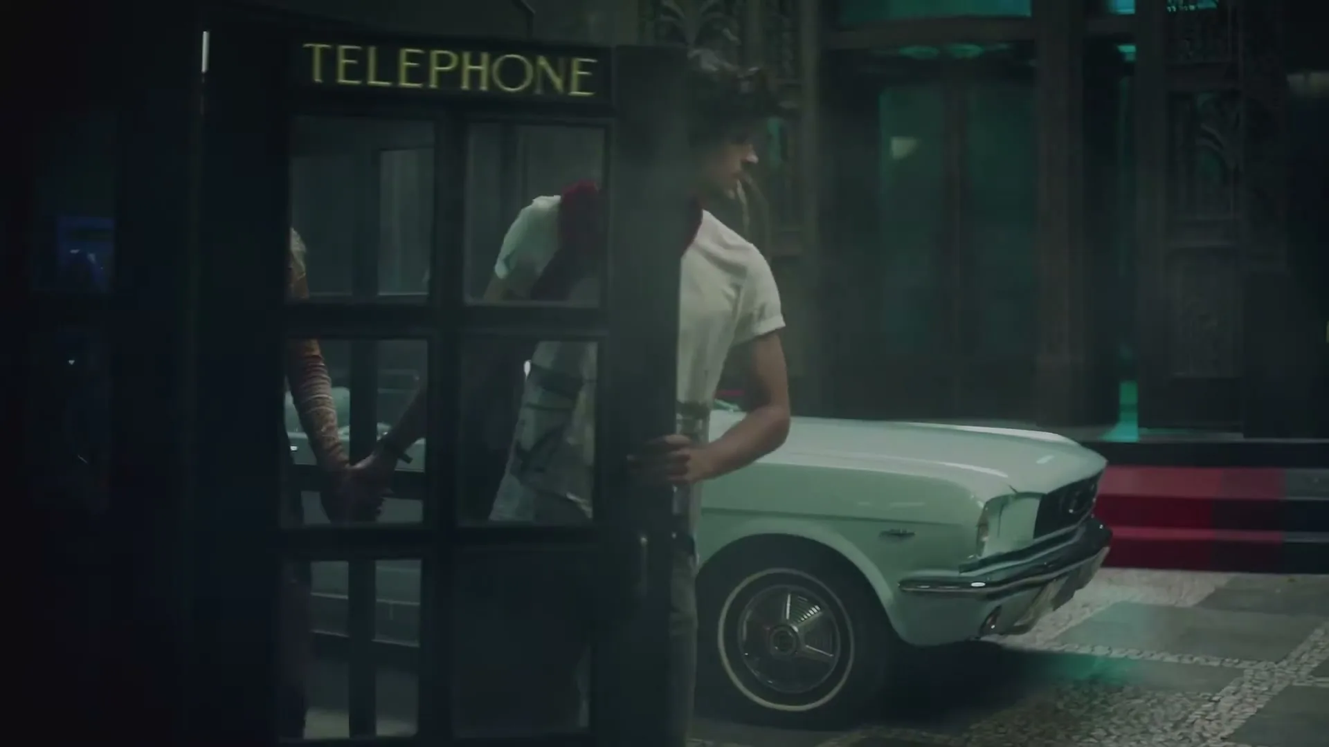 A young couple, a man and a woman, holding hands and walking towards a telephone booth, with a vintage car parked in the background.