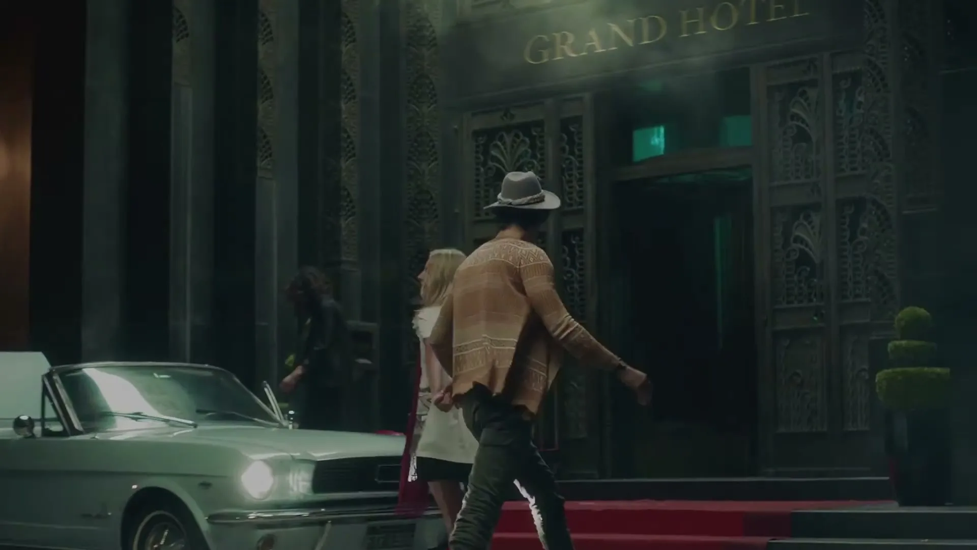 A man in a hat and a woman in a white dress walk towards the entrance of a grand hotel, with a classic car parked nearby.