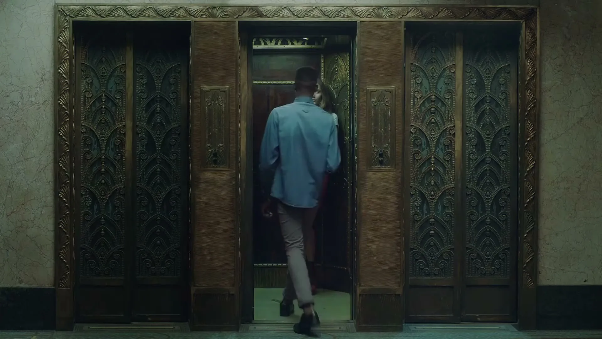 A young man in a blue shirt walks into an ornate elevator where a young woman in a white shirt and pink skirt is standing, in a dimly lit building.