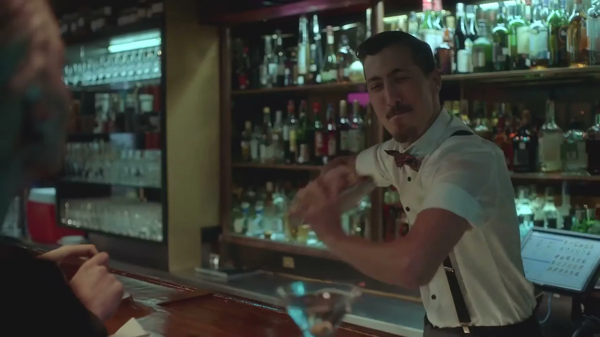 A male bartender with a mustache and bow tie shakes a cocktail shaker behind a bar, with shelves of liquor bottles in the background.