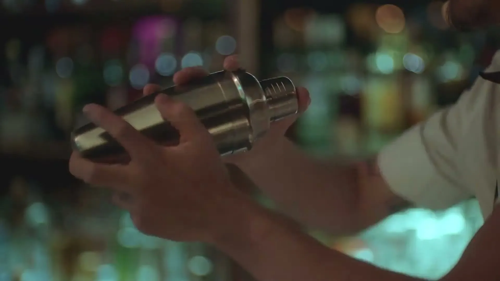 A bartender's hands shake a cocktail shaker in a dimly lit bar with blurred bottles in the background.