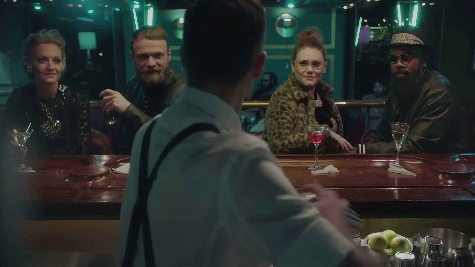 A bartender in a white shirt and suspenders mixes a drink for four diverse patrons sitting at a dimly lit bar.