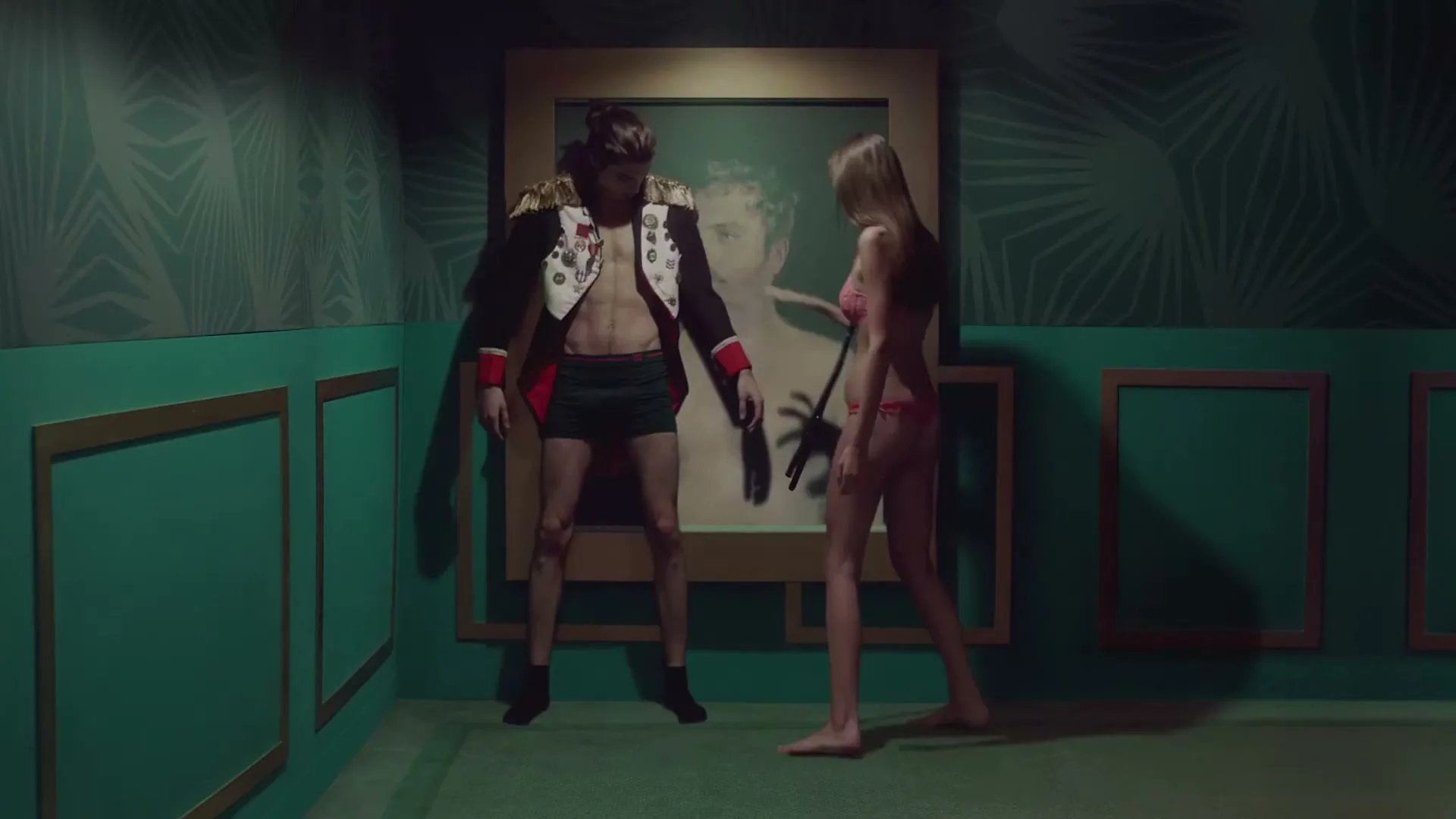 A man and a woman in underwear stand in a green room, as the man's outfit and the painting behind them change.