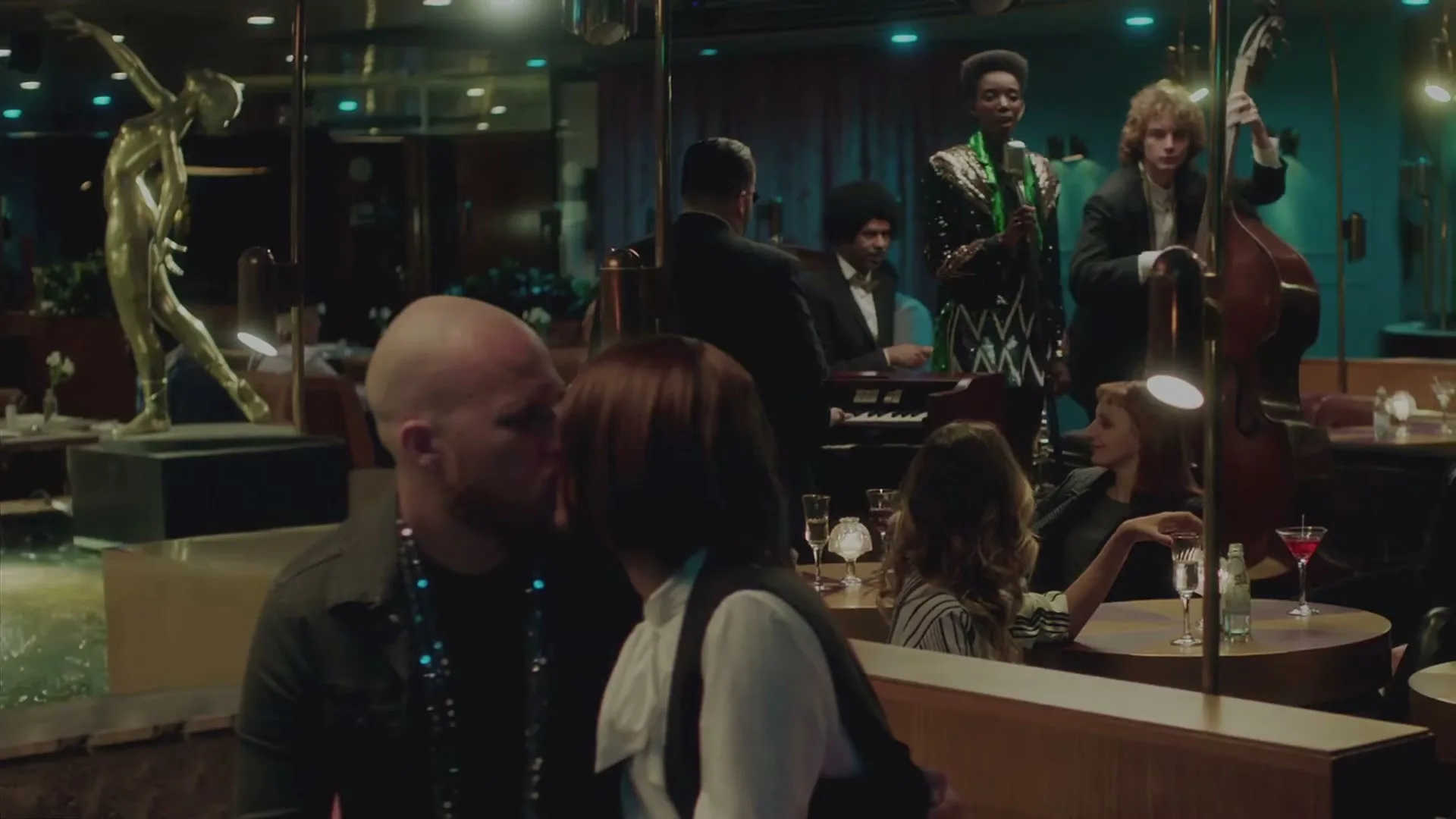 A bald man and a woman with a bob haircut kiss in a dimly lit restaurant with a band playing in the background.