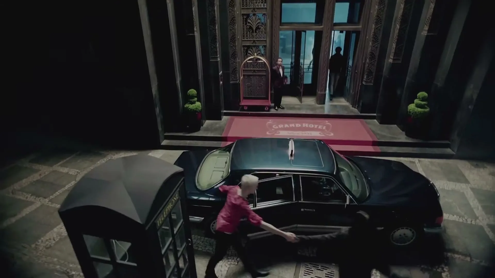 A blonde woman in a red shirt exits a black car, shaking hands with a dark-haired man, outside a grand hotel entrance at night.