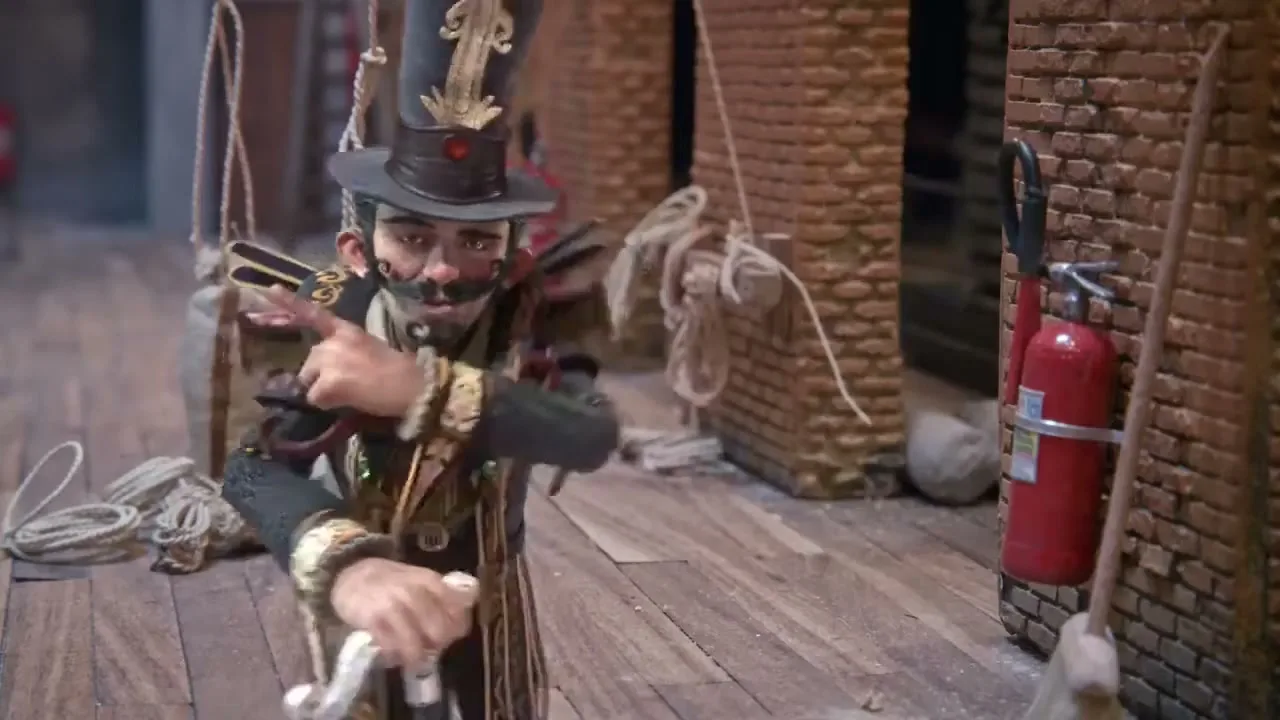 A stop-motion puppet, dressed as a ringmaster, gestures dramatically while a sign with "APLAUSO" (applause) appears, in a backstage setting.