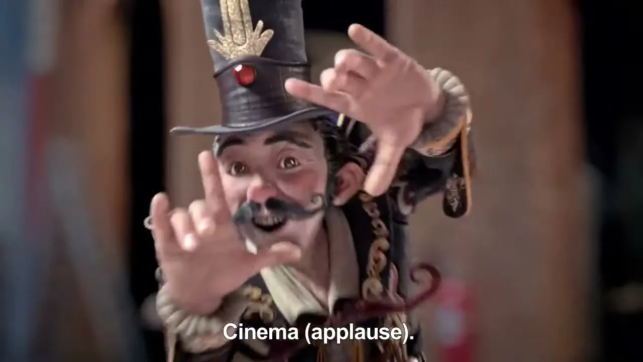 A stop-motion puppet with a top hat and elaborate costume gestures with its hands, framing its face, in a theatrical setting.