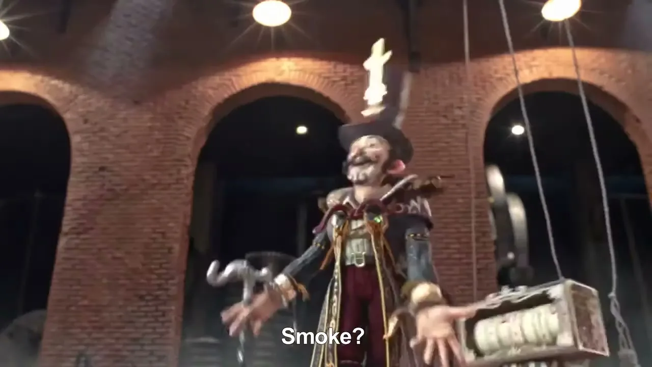 A male character with a painted face and a top hat, wearing an elaborate costume, stands with arms outstretched in a brick-walled industrial setting.