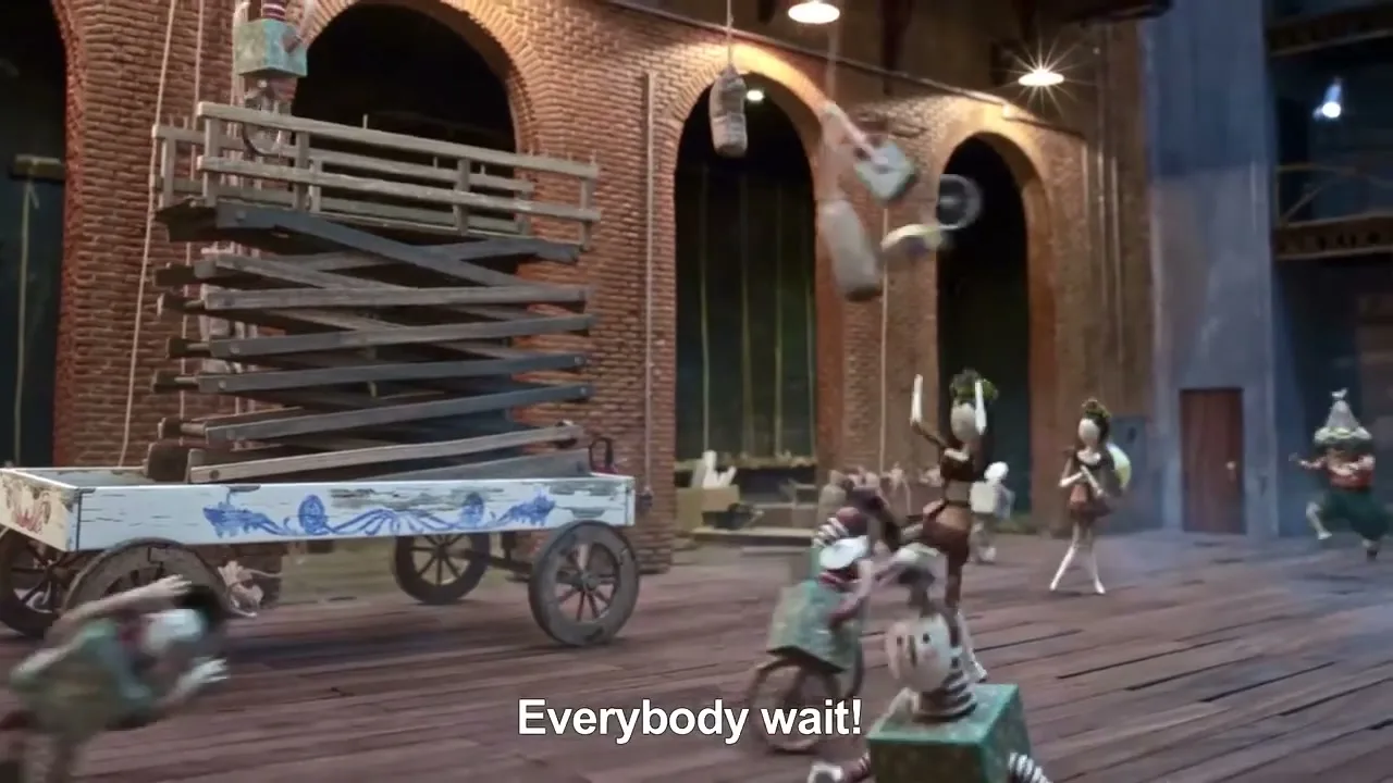 A group of animated wooden puppets, some on bicycles, others running, move chaotically around a large wooden cart on a stage with brick arches in the background.