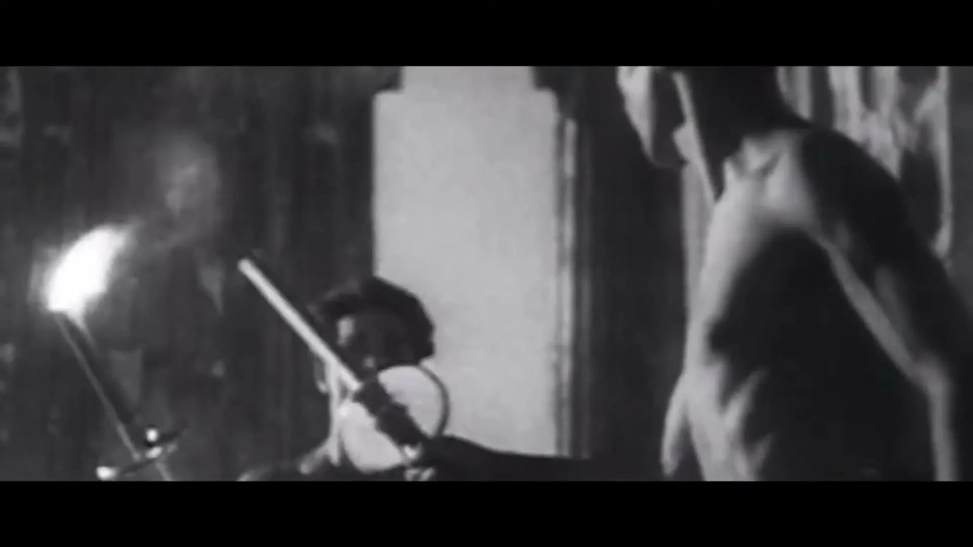 Two men, one shirtless, practice fencing with swords and a torch in a dimly lit, old-fashioned room.