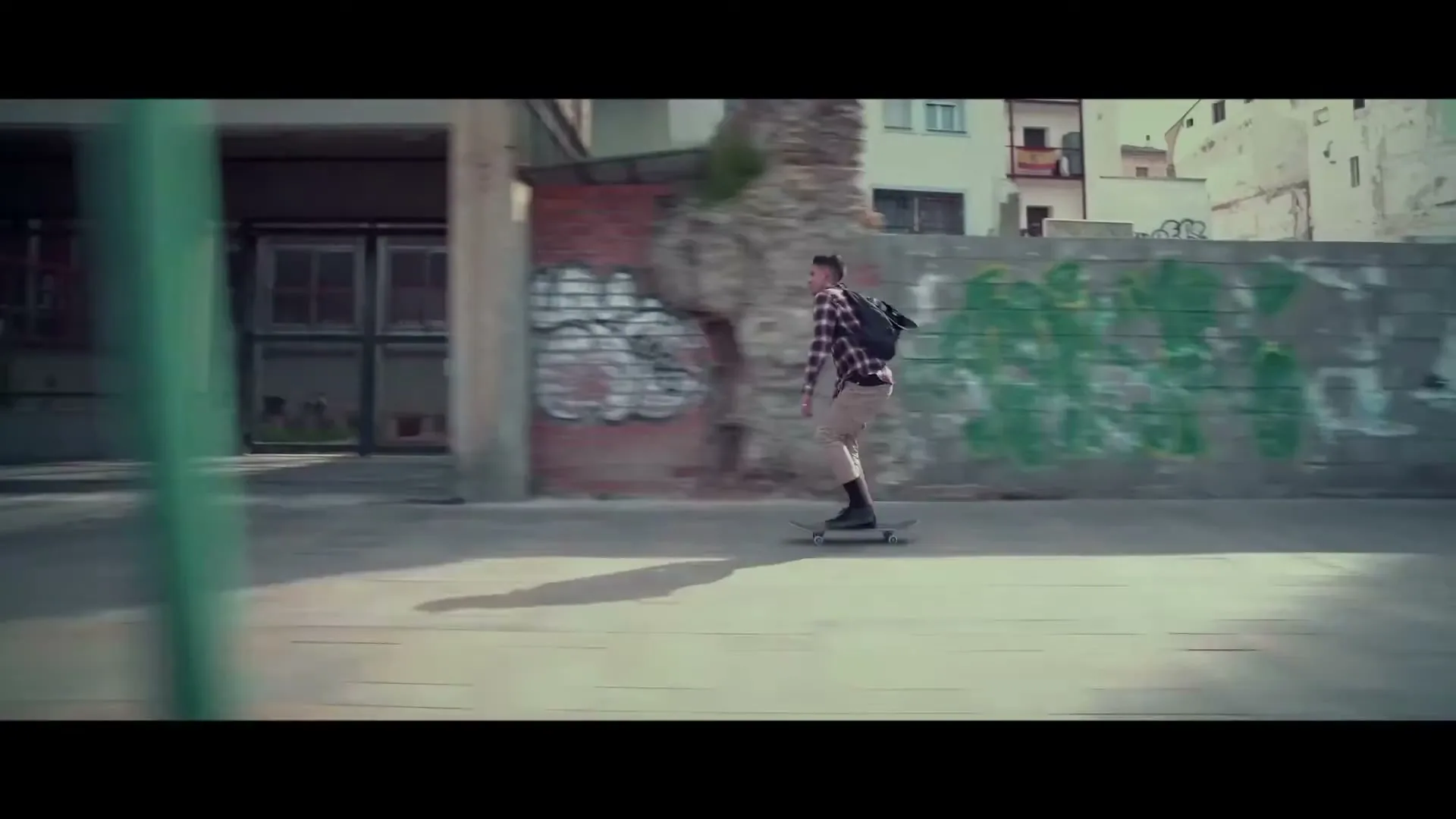 A young man with a backpack and plaid shirt skateboards down a street past a building with graffiti on a sunny day.