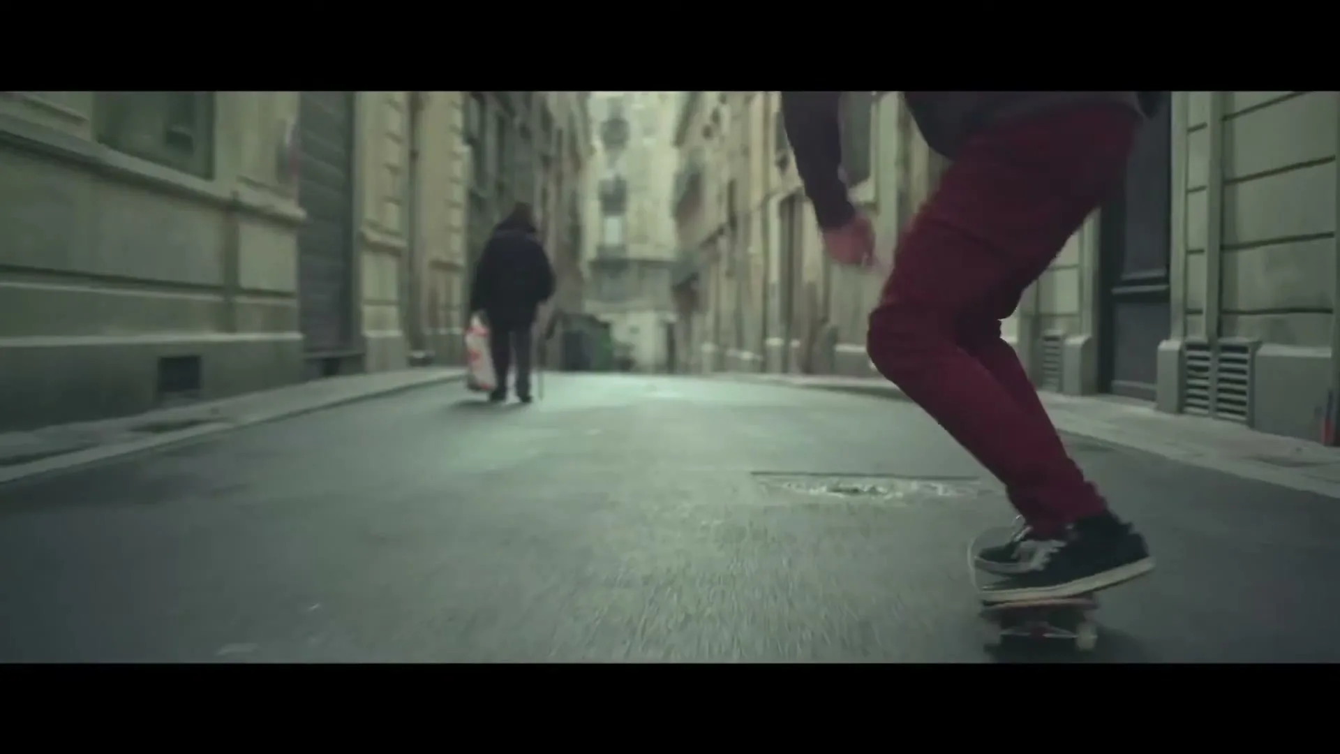 A person in red pants and black shoes rides a skateboard down a narrow, old city street, with another person walking in the distance.