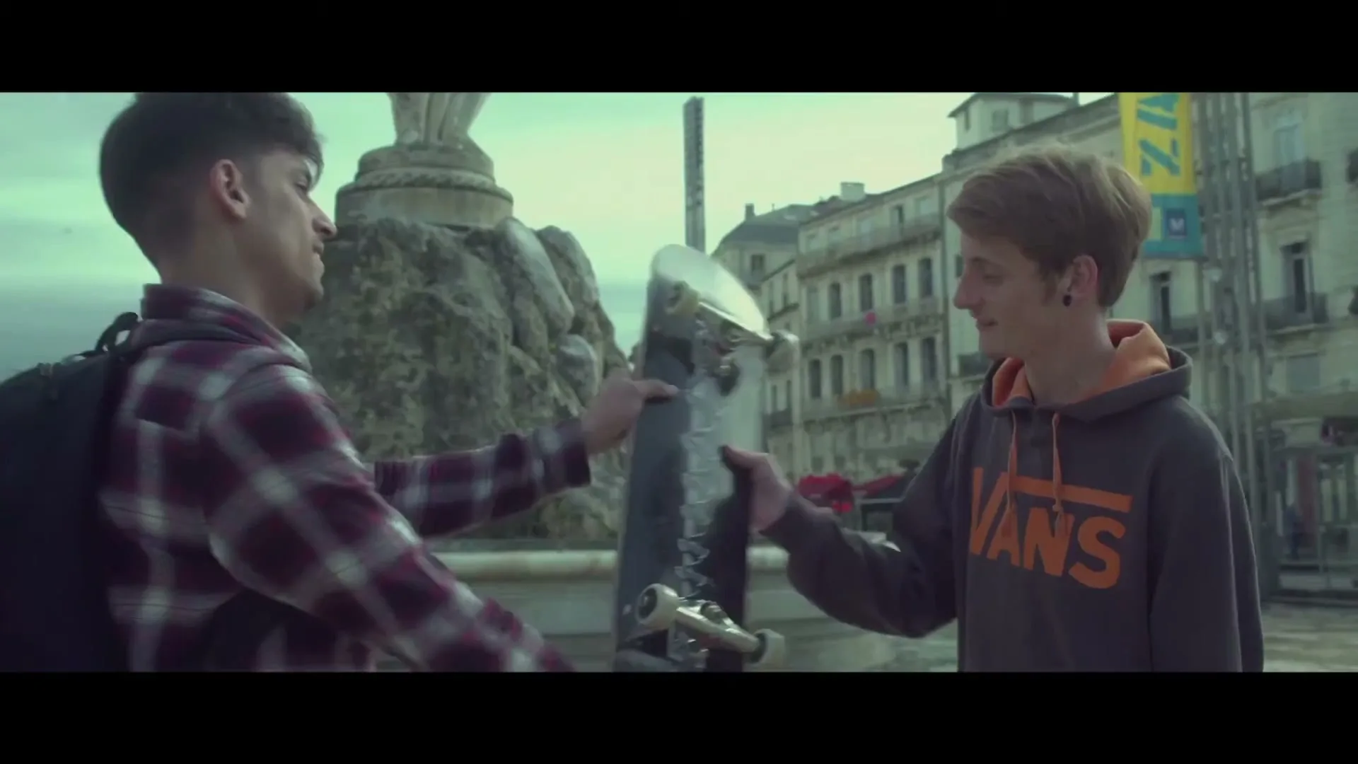 Two young men, one in a plaid shirt and the other in a Vans hoodie, exchange a skateboard in a European city square.