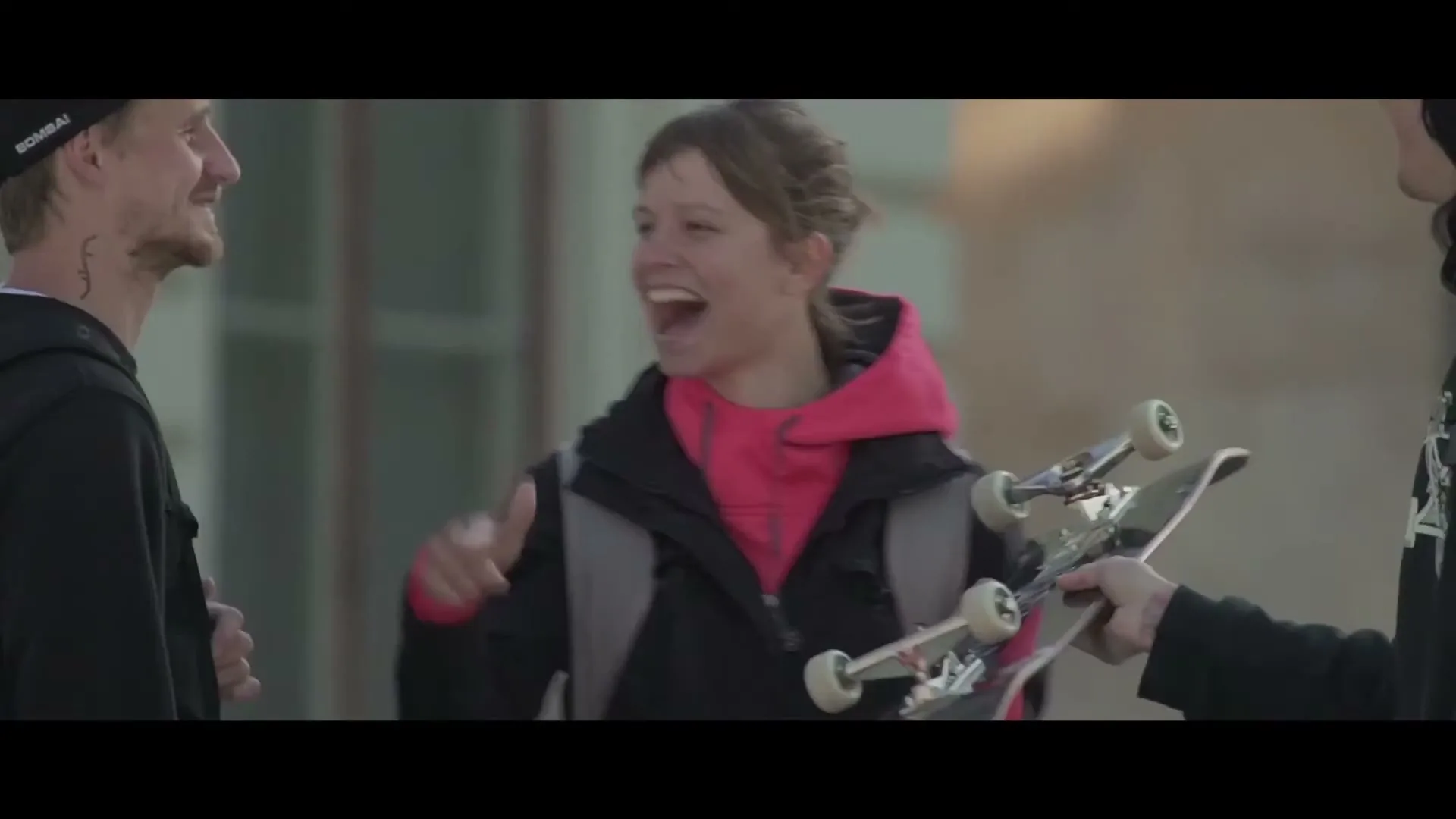 A young woman in a black jacket and pink hoodie laughs with two men, one holding a skateboard, outdoors during the day.
