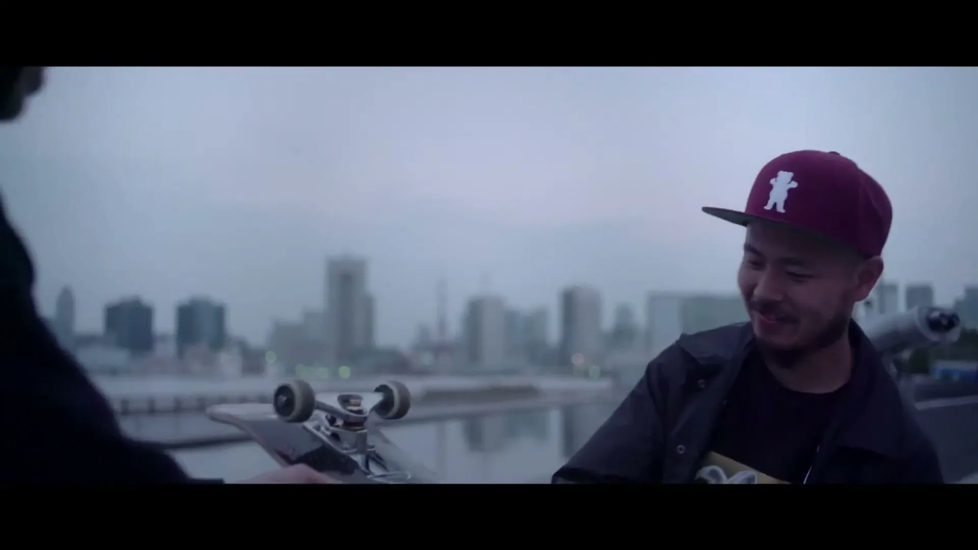 A young Asian man in a cap and jacket holds a skateboard, smiling and looking at someone off-screen, with a city skyline in the background.