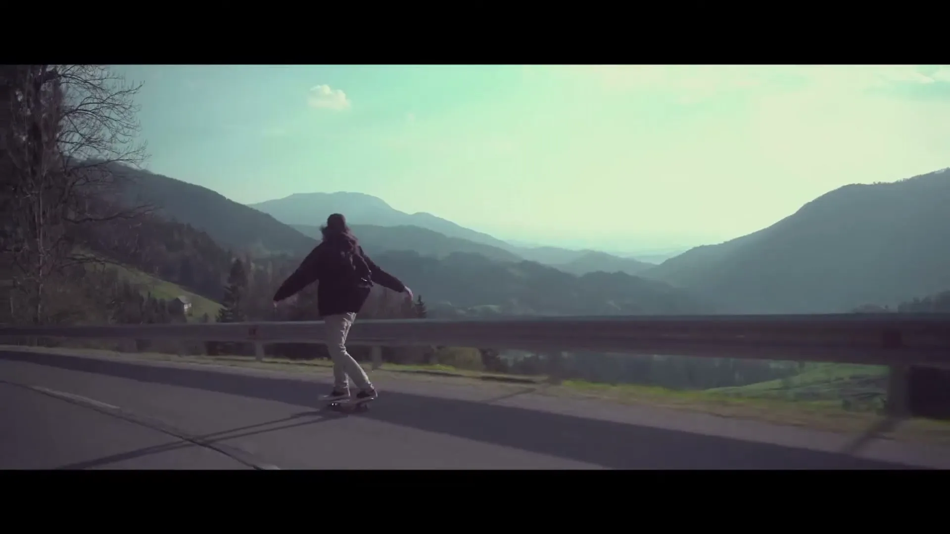 A person with long hair and a backpack skateboards down a winding road, with mountains and trees in the background under a bright sky.