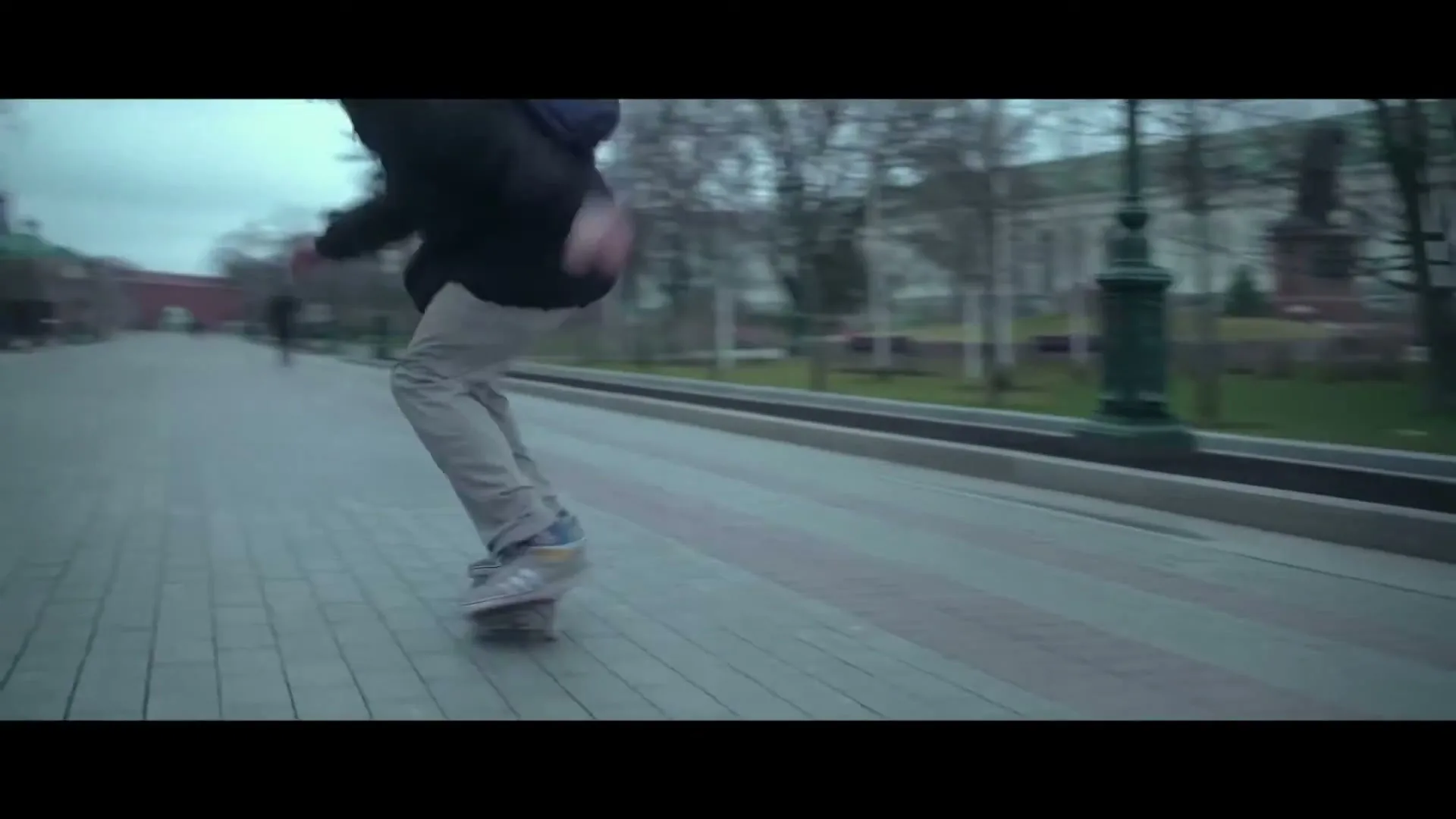 A male skateboarder in a dark jacket and light pants performs a trick on a paved path in a park on an overcast day.