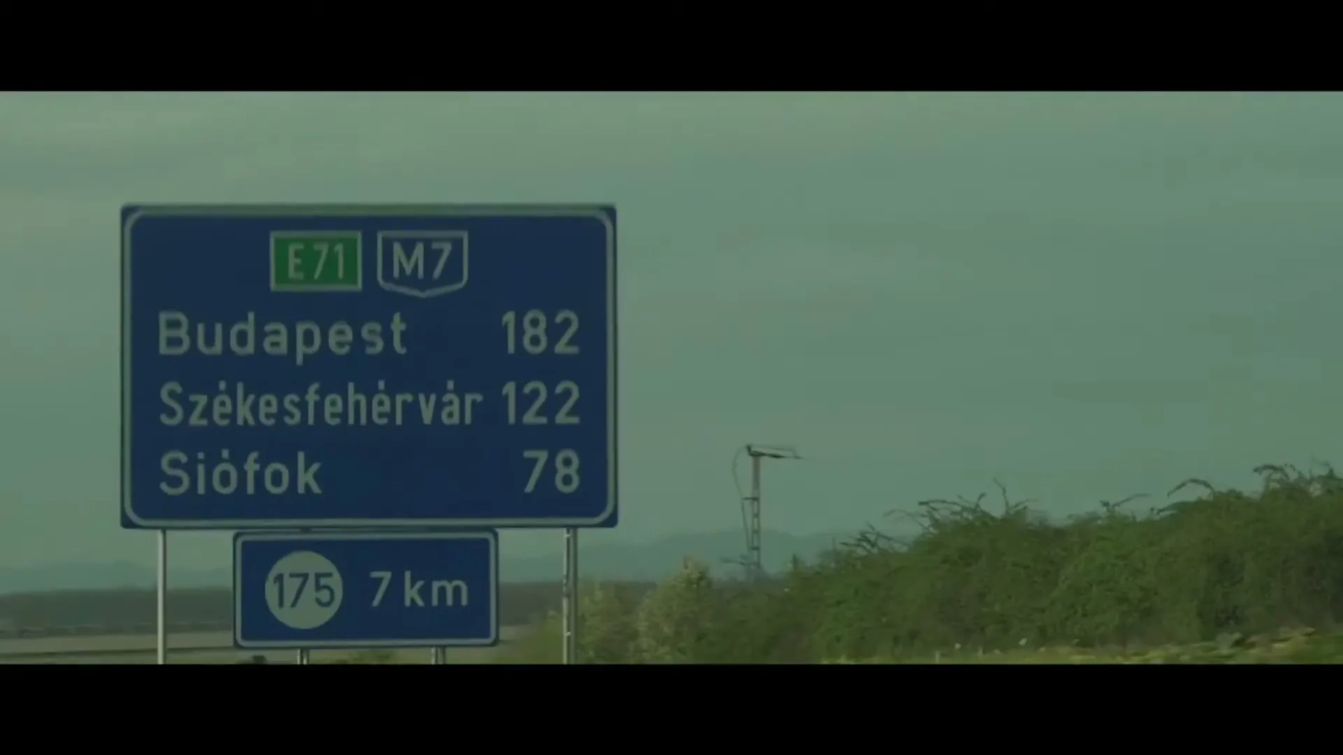 A blue road sign with white text indicating distances to cities like Budapest, Sz
r
ekesfeh
r
v
r and Si
r
fok stands by a highway under a cloudy sky.
