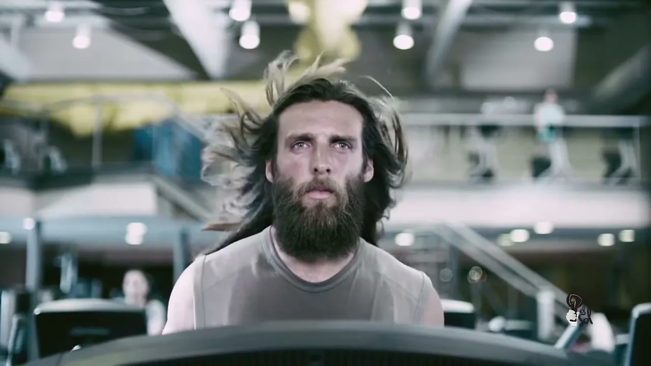 A man with long hair and a beard runs on a treadmill in a gym, looking tired and sweaty.