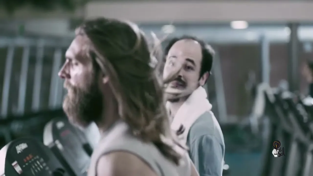 A man with long hair and a beard runs on a treadmill, his hair flowing behind him, while another man with a goatee watches him with envy in a gym.