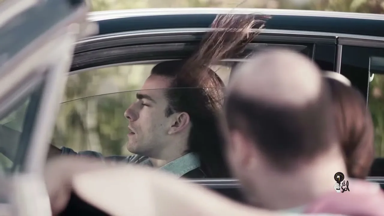 A man with long hair drives a car, his hair blowing in the wind, while a bald man is seen in the foreground.