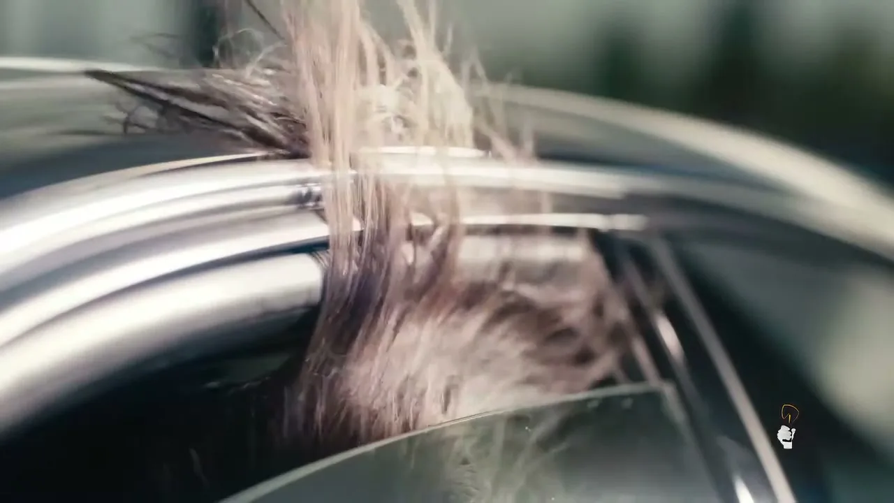 A close-up shot shows a woman's long, dark hair flowing out of a car window as she drives, with sunlight catching the strands.
