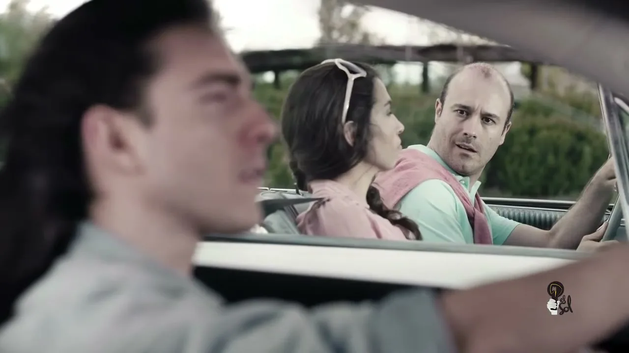 A man with long hair drives a convertible while a couple in the back seat looks at him with envy on a sunny day.