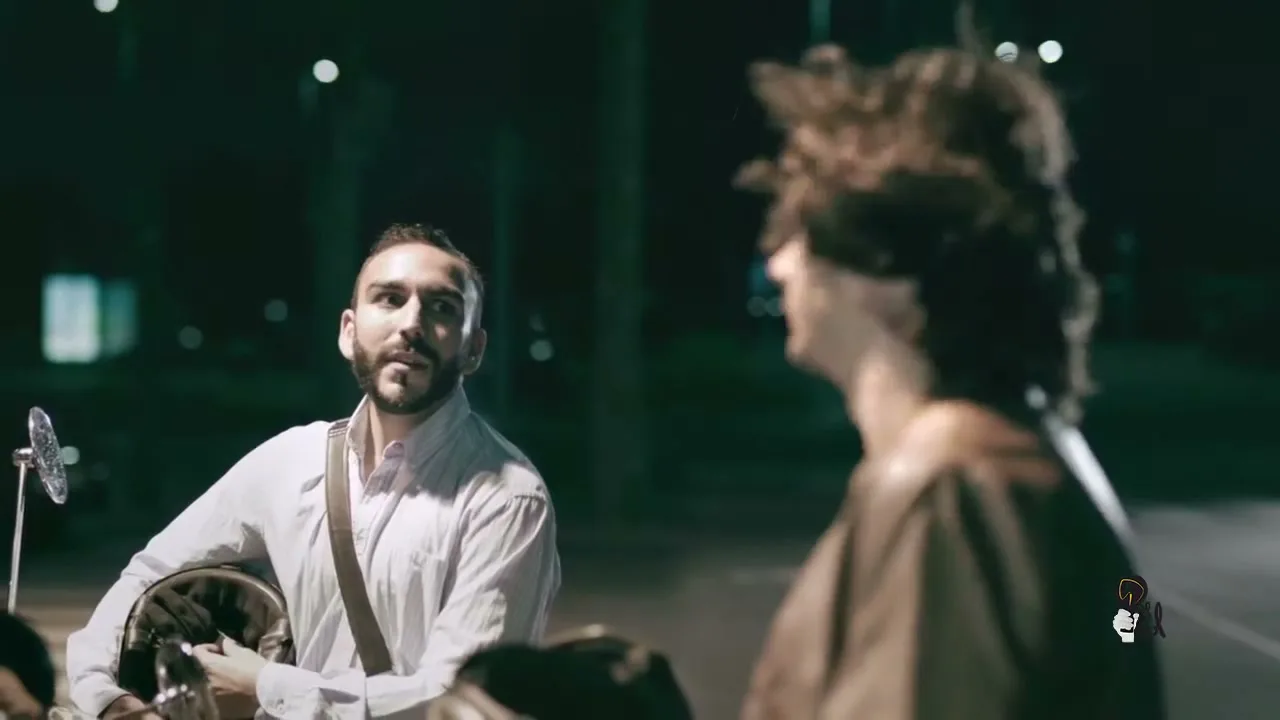 Two men are talking at night, one with a beard and white shirt, the other with curly hair and a brown jacket, on a dark street.