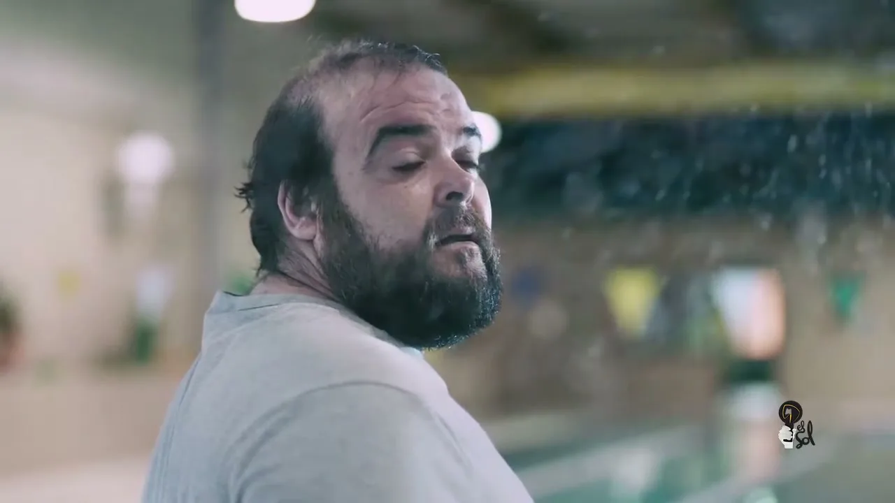 A balding, bearded man in his 40s, wearing a grey t-shirt, looks over his shoulder in a swimming pool, as water splashes around him.