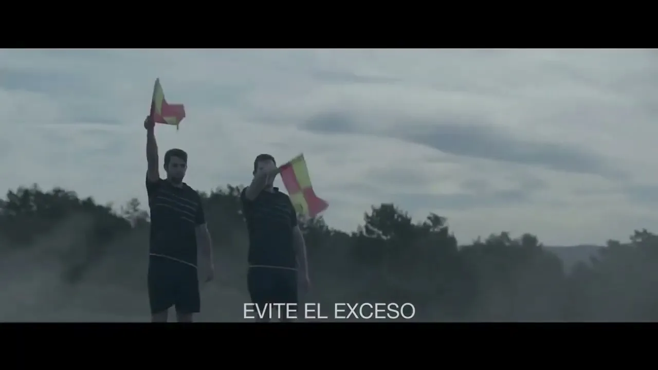 Two male soccer line judges in black uniforms stand on a dusty field, raising their red and yellow flags under a cloudy sky.