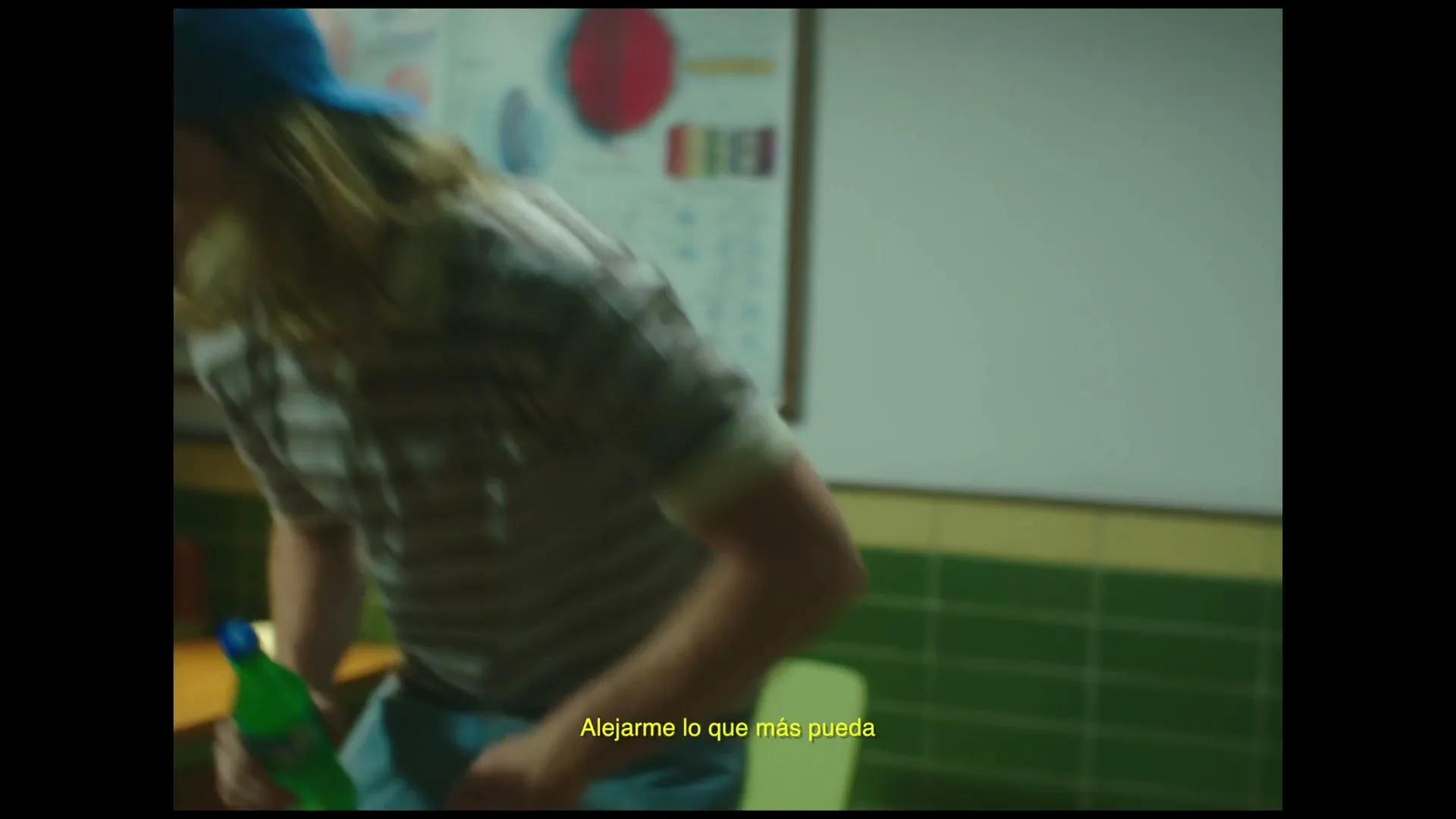 A young man with long blonde hair and a blue cap enters a classroom, holding a Sprite bottle, and sits down at a desk.