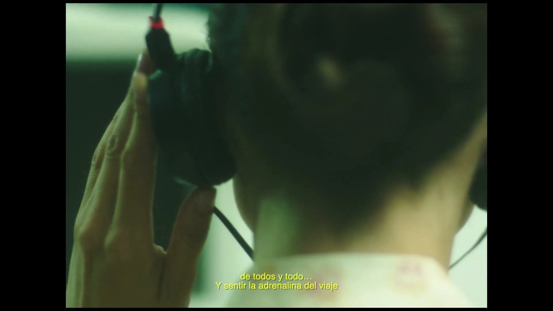 A person with short hair and a patterned shirt adjusts headphones, seen from behind in a dimly lit interior.