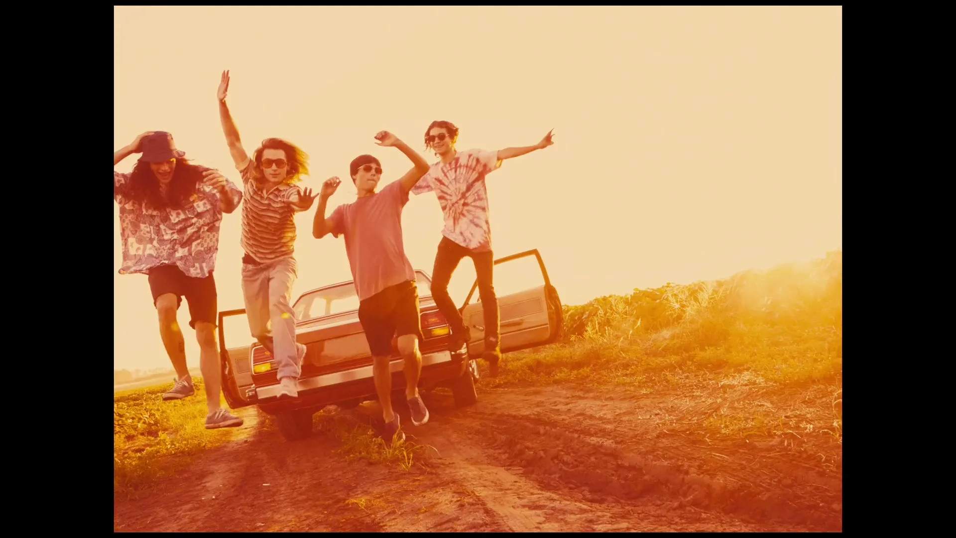 Four young men in casual clothes jump out of a car on a dirt road in a field at sunset, looking joyful.