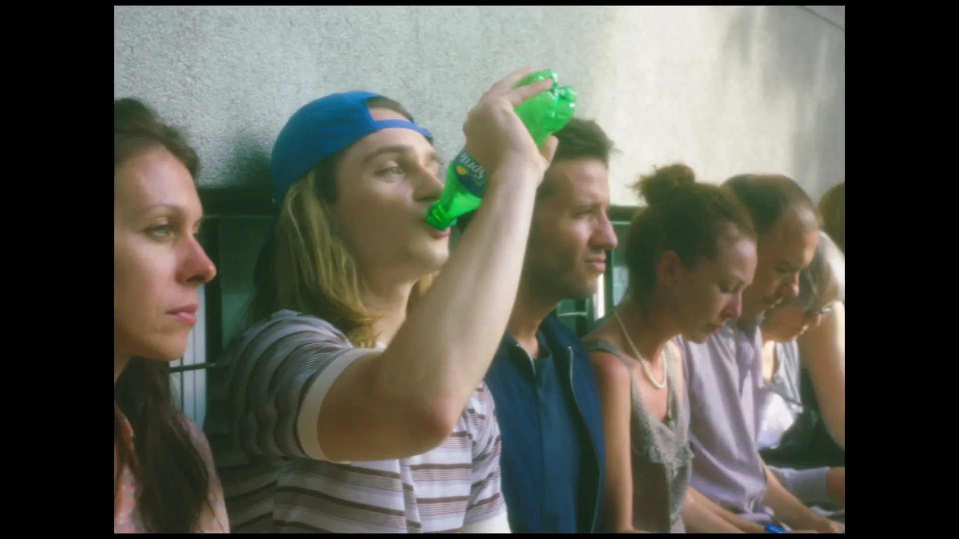 A young man with long blonde hair and a blue cap drinks from a green Sprite bottle while sitting on a bench with other people.