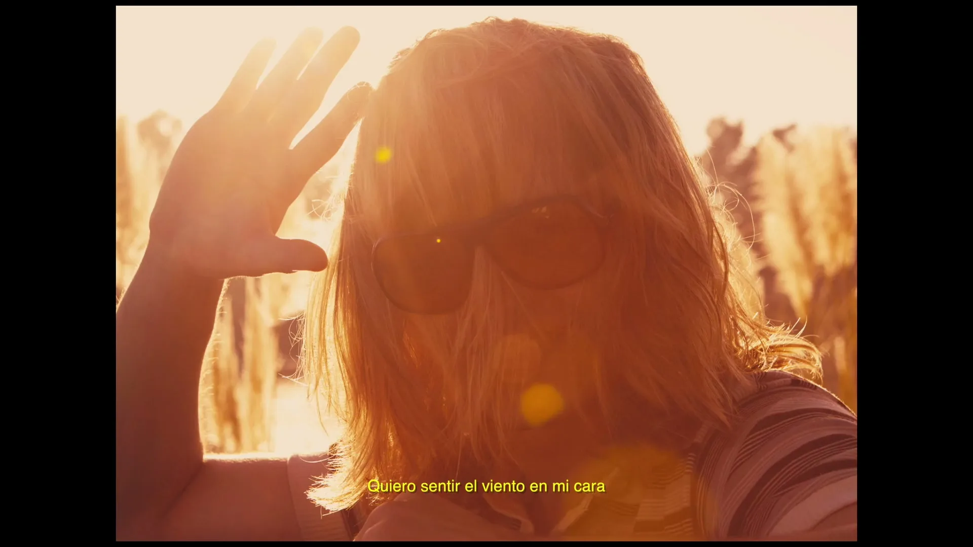 A person with long blonde hair and sunglasses shields their eyes from the bright sun in a field of tall grass during golden hour.