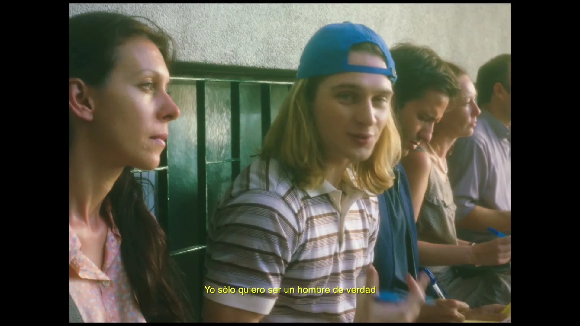A young man with long blonde hair and a blue cap sits in a row of people, looking forward and speaking, in an interior setting.