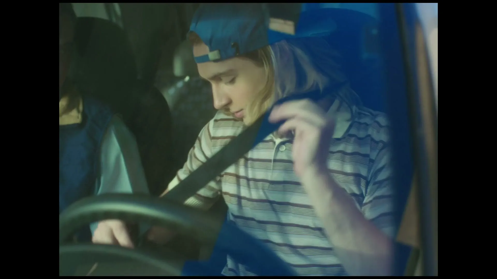A young man with long blonde hair and a blue cap puts on his seatbelt while sitting in the driver's seat of a car at night.