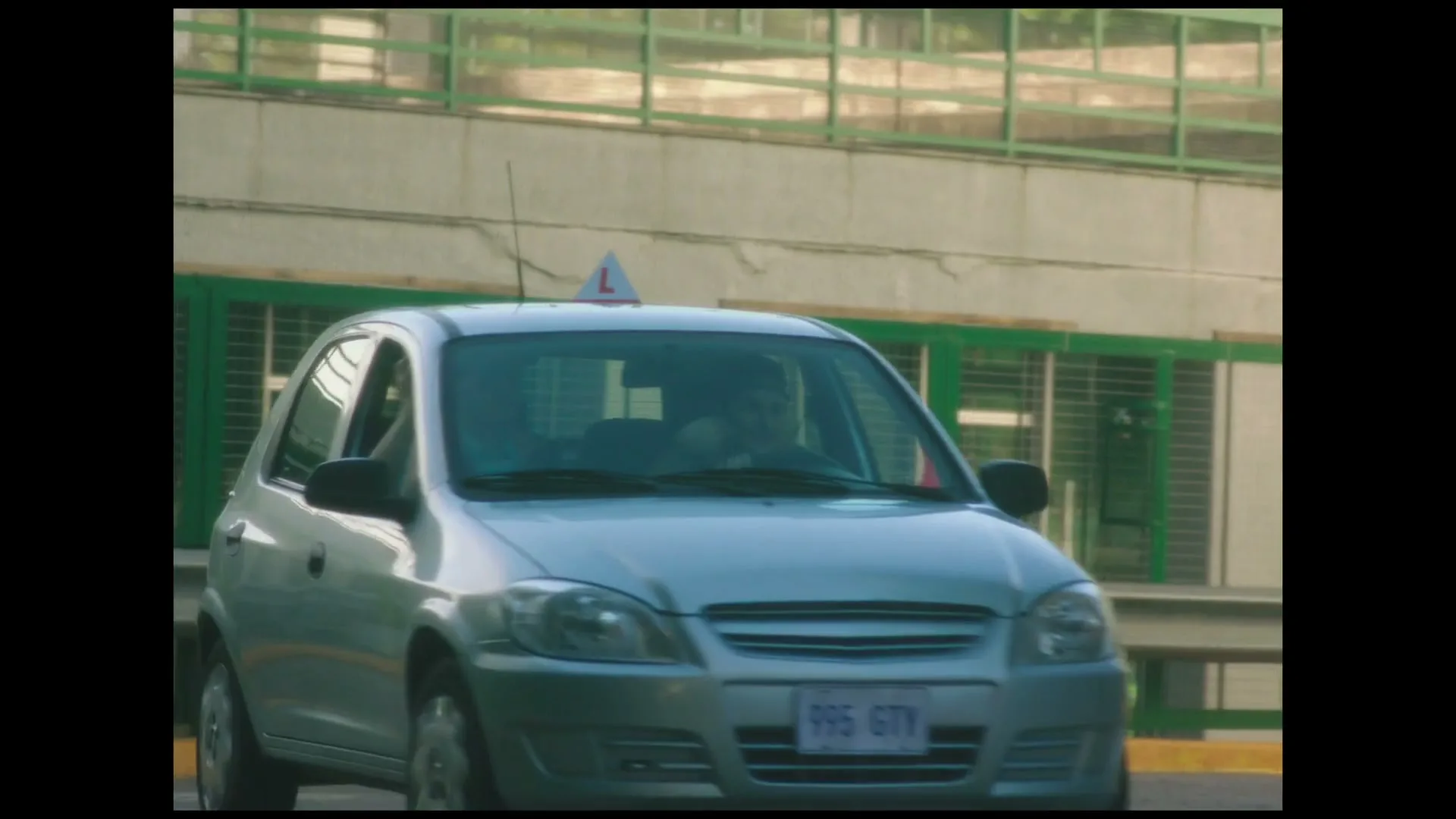 A silver car with a "learner" sign on top drives past a red traffic cone and a green fence on a sunny day.