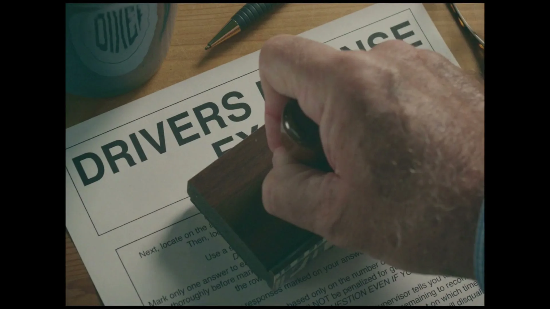A male hand stamps a "DRIVERS EXAM" paper with a red "FAILED" stamp on a wooden desk, next to a pen and a mug.