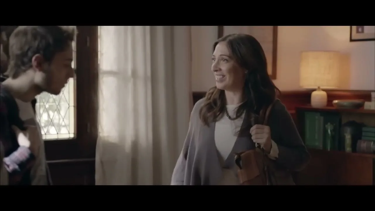 A woman with long brown hair and a grey cardigan enters a room, smiling and looking at a young man who is partially visible on the left.
