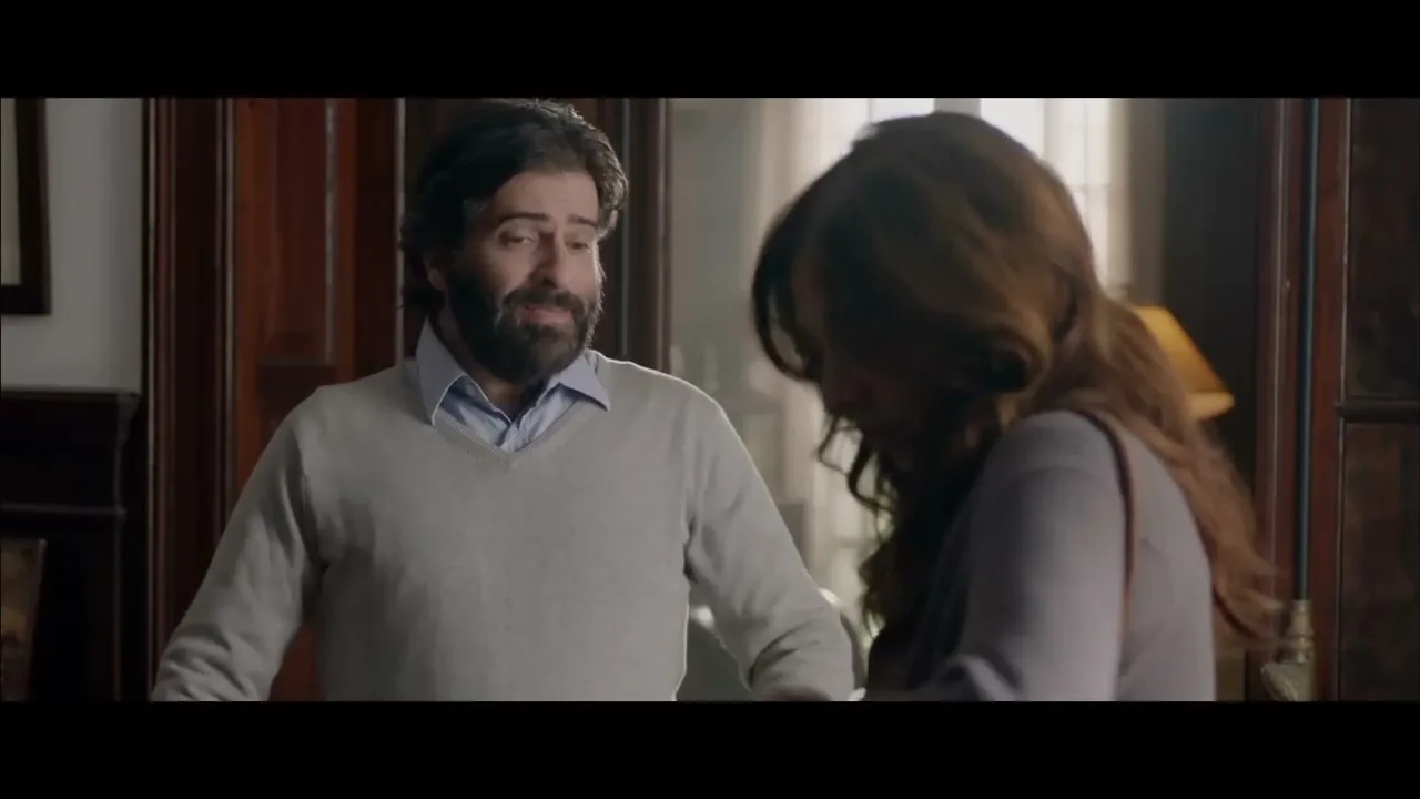 A bearded man in a light sweater and a woman with long brown hair talk in a dimly lit room with dark wood furniture.