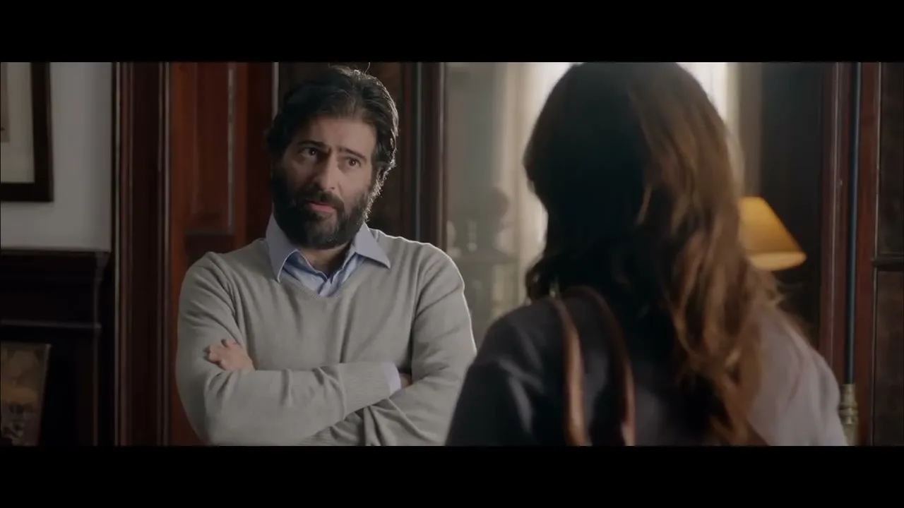A bearded man in a light sweater and blue shirt stands with crossed arms, looking at a woman with long brown hair from behind in a dimly lit room.