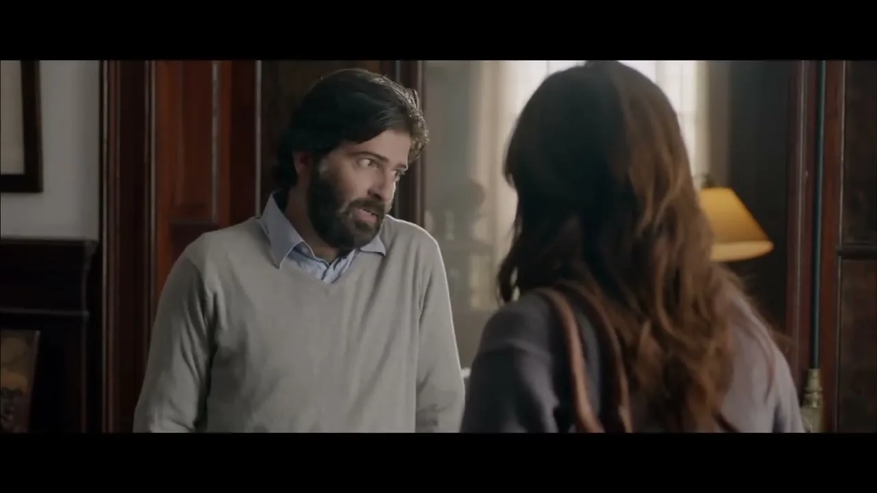 A male therapist with a beard talks to a female patient from behind, who is holding a purse, in a dimly lit office.