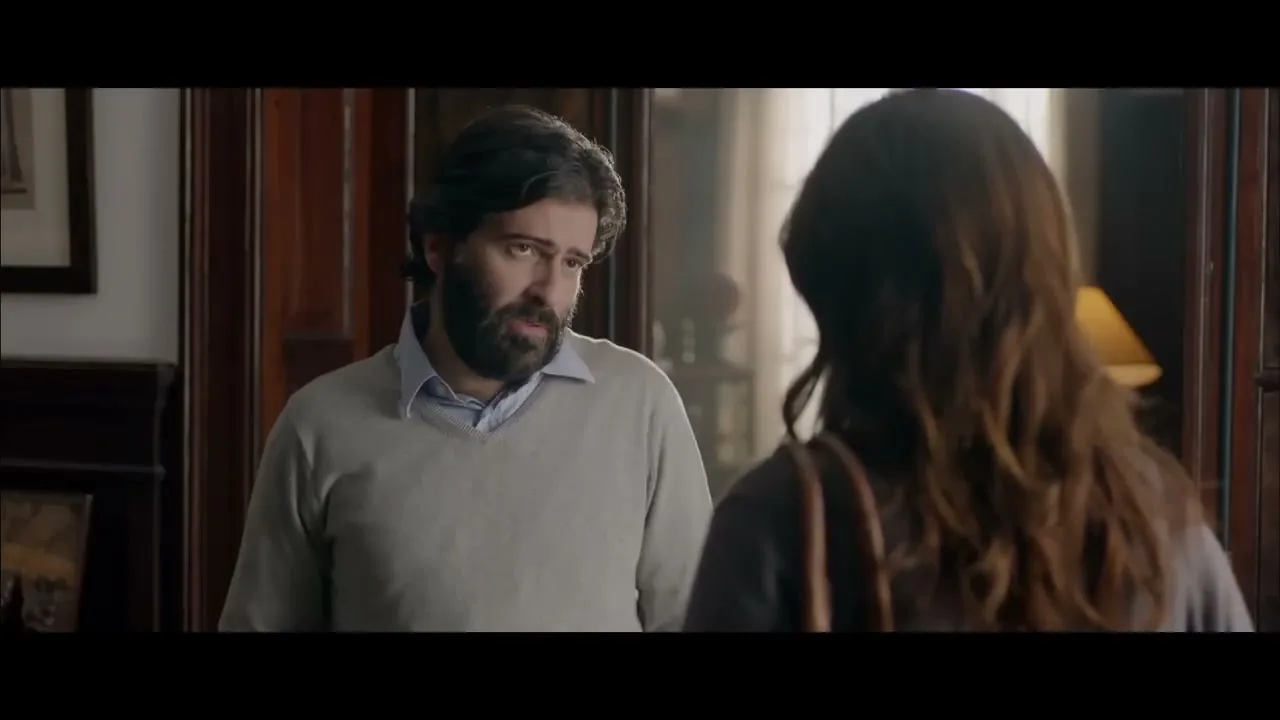 A male therapist with a beard and a light sweater talks to a female patient from behind in a dimly lit office.