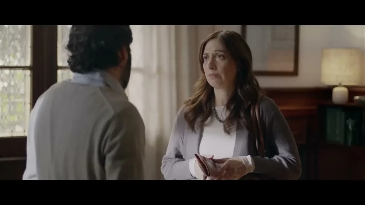 A woman with long brown hair, wearing a white shirt and grey cardigan, holds a phone and looks at a man in a therapist's office.