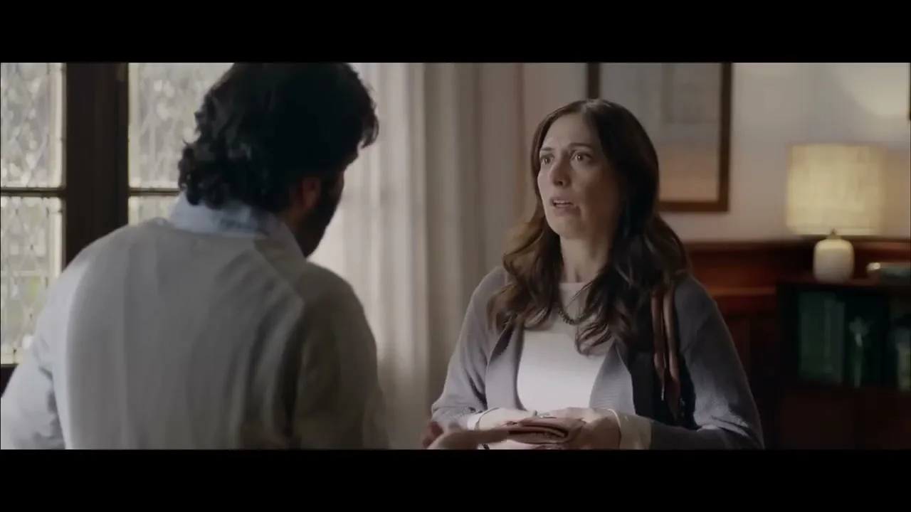 A woman in her 40s, wearing a grey cardigan, talks to a man with his back to the camera in a dimly lit office.
